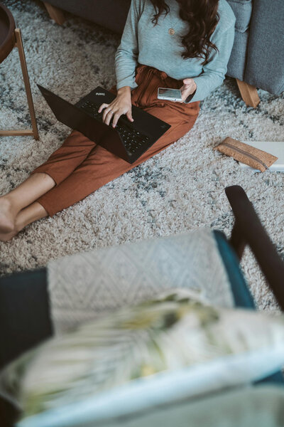 In a cozy, modern home setting, a student is seated on the floor with a laptop on their lap and a smartphone in hand.
