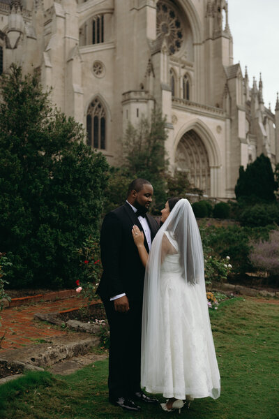 A wedding couple, the groom is a Black guy wearing a tux and the bride an Asian and Mexican woman wearing an ivory color vintage dress with a veil. Both are looking at each other in love in a garden in front of the National Cathedral.