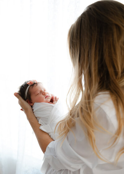 Mom holds and looks down at hernewborn baby girl wrapped in white swaddle.