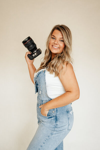 Portrait of Taylor Ragusett, a smiling Charleston photographer holding a camera, wearing denim overalls.