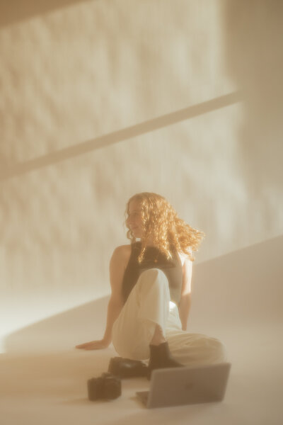 The wedding photographer, Autumn, sitting on the floor of an off white backdrop during golden hour.