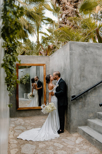 Bride and groom kiss in a modern courtyard with concrete walls, lush palms, and a reflection in the mirror at Acre Resort in Mexico.