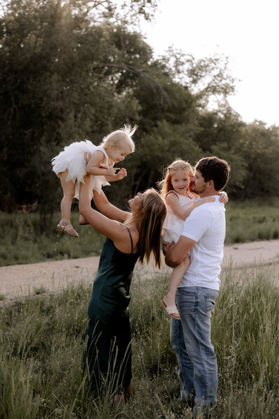 Gorgeous family playing with their children in a field of tall green crass with a row of trees in the background. Mary Wiebe Photography  took this image.