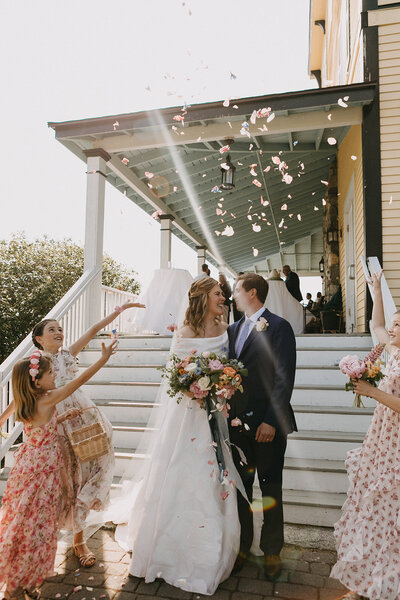 New England couple smiling at each other as flower girls throw petals around them