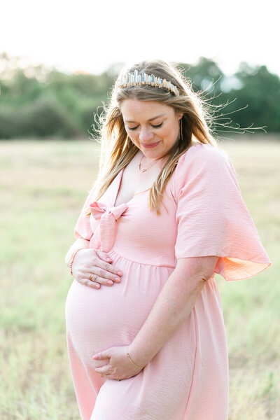 Cedar Park Photographer | A pregnant woman dressed in a pale pink dress stands in a field of tall grass at Brushy Creek Lake Park. She looks down at her belly with a soft smile of her face in this tender shot.