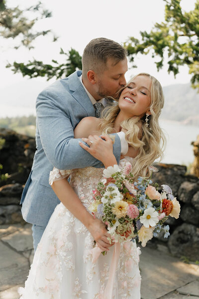 A posed wedding photo of a couple at the Griffin House wedding venue in Hood River, Oregon. The couple is standing in the center of the photo  embracing each other. There are stone pavers under them, a rock wall behind them, and a view overlooking the Columbia River Gorge. Their wedding ceremony flowers are on the ground and made up of colorful flowers in pink, light purple, light blue, and soft yellow. The bride is holding her fresh floral bouquet. The groom has his hand in his pocket. The bride is wearing a white  dress with a long train. The groom is wearing a powder blue suit.