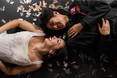 LGBTQ+ couple in a wedding gown and suit lying on the ground laughing together surrounded by confetti