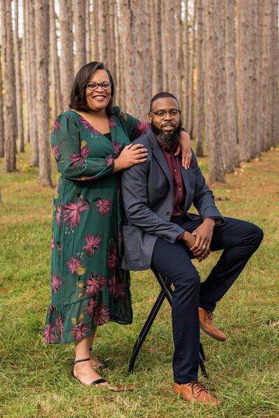 a mom and ad pose together. The dad sits on a stool, wearing a blue jacket and the woman wears a teal floral dress