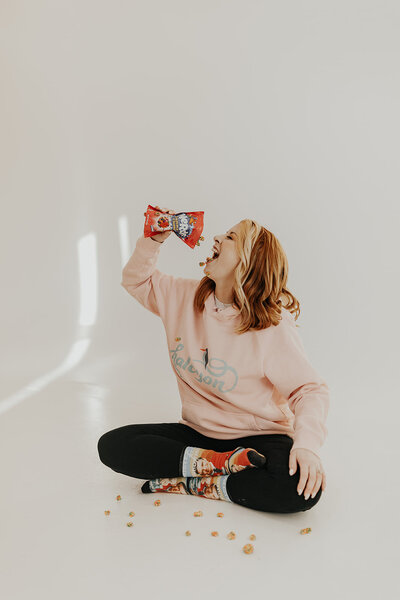 Shylah sits on the floor, cross-legged, wearing Mr. Rogers' socks and a pink sweatshirt with the Halcyon logo. She is pouring Nerds candy clusters into her mouth, several of which have fallen on the floor around her feet.