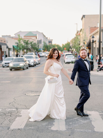 Bride and groom holding hands and crossing a busy street, smiling at the camera.