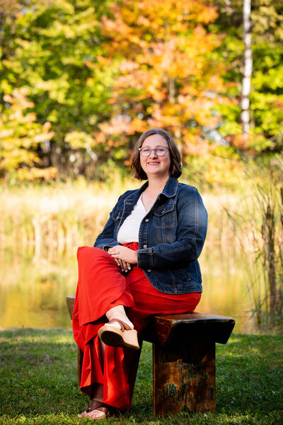 Kasey Pierce, wearing rust colored pants, a white blouse, denim jacket, and glasses, sits on a brown bench outside in front of a background of autumn trees
