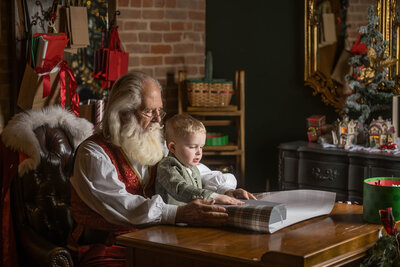 Older man with a white beard reads a book to three children in red dresses by a table. Christmas trees and stockings set a festive atmosphere.