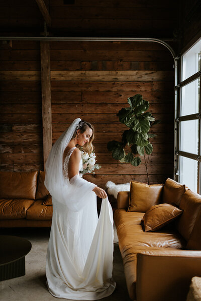 The bride, in a white gown and veil, stands indoors holding her dress beside a tan sofa and a large potted plant before her wedding at 100 Acre Wood in Picton, ON. The scene, captured by Toronto wedding photographer, Jennifer van Son.