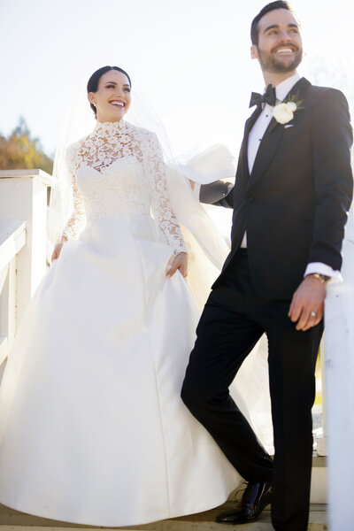 Bride in a high-neck lace wedding gown and groom in a classic tuxedo walk together in the golden sunlight at Omni Bedford Springs Resort, smiling joyfully.