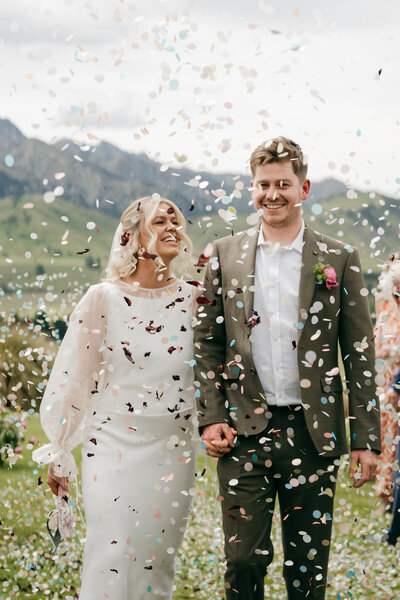 Bride and groom smiling and holding hands as flower petal confetti falls around them, with a scenic mountain landscape in the background. Wedding Photography by Melissa Clark