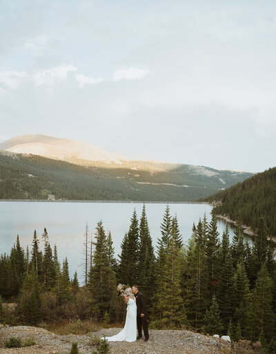 bride and groom are kissing as the sun sets. there are mountains behind them in colorado with snow on them. the trees are also in the background of the overlook.