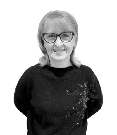  Smiling Salon Co-Ordinator with shoulder-length hair, glasses, and a black sweater featuring subtle embellishments, showcasing professionalism and warmth.