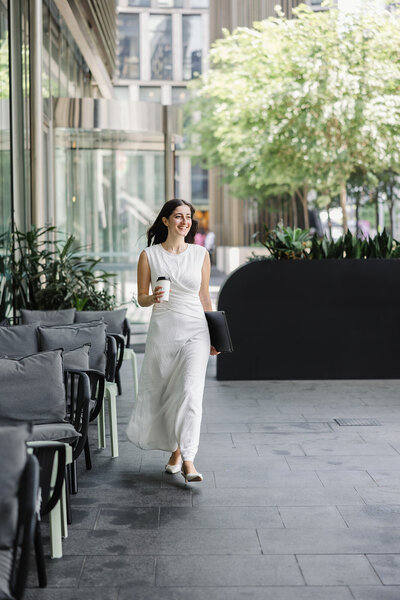 Woman in a white dress walking confidently with a coffee cup and laptop in hand through a modern Sydney business district, captured during a personal branding session by Karen Sadek.