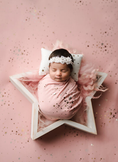 A tiny little newborn baby girl smiles as she's carefully posed and swaddled in a photography prop.