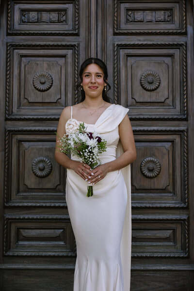 Portrait of the bride, Katie, holding her bouquet standing in front of a door.