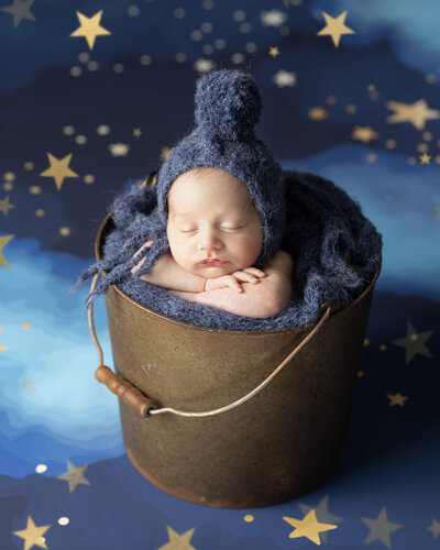 Sleeping newborn boy in gold bucket, wearing navy blue bonnet, on night sky backdrop with gold stars
