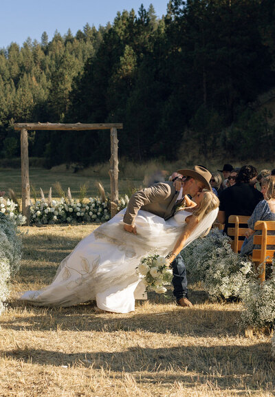 Groom in a cowboy hat dipping the bride for a kiss under their ceremony arbor during an outdoor wedding on private property.