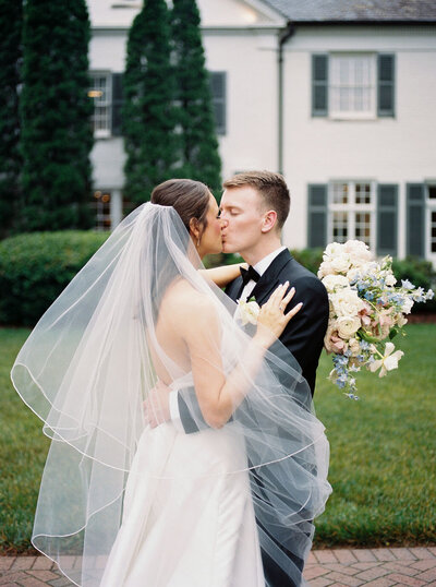 Seth and Mimi share a passionate kiss on the grounds of Boxwood Estate, surrounded by lush greenery and scenic landscaping. Mimi wears a stunning white wedding gown, and Seth is dressed in a classic suit. The soft, natural light adds a warm, romantic glow to the moment, capturing their love and joy in a picturesque setting.