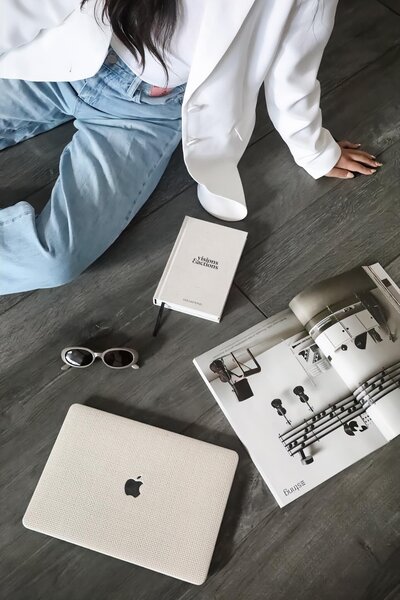 Flat lay of Jenny  sitting on a wooden floor with a MacBook, sunglasses, a notebook, and an open magazine, showcasing a minimalist and stylish work setup