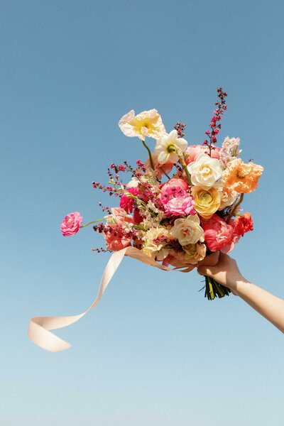 Bright spring elopement bouquet by BLOOM BLOOM with ribbon blowing in the wind against a blue sky