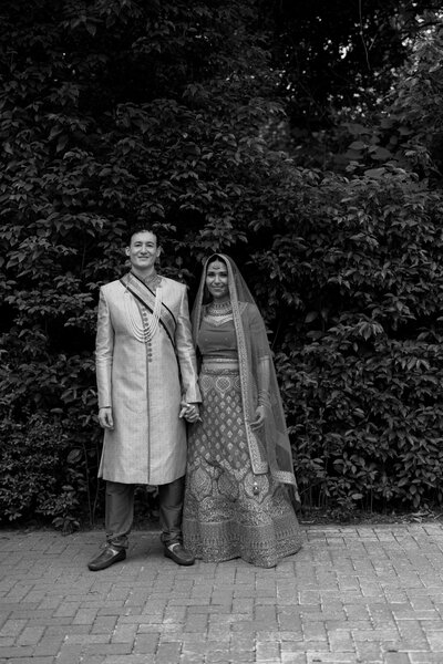 April M Payne Photography captured a couple dressed in traditional Indian wedding attire, standing hand in hand in front of lush greenery, with the bride wearing a colorful lehenga and veil, and the groom in a beige sherwani.