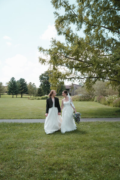 LGBTQ+ couple in their wedding dress holding hands and looking at each other as hey walk towards the camera in a lush green field in the Berkshires in Massachusettes.