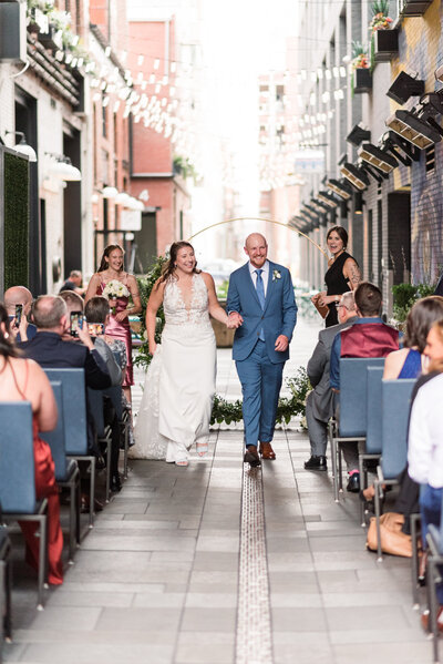 urban wedding in downtown denver with bride adn groom holding hands and walking down the aisle together after their ceremony with twinkle lights overhead captured by denver photographer