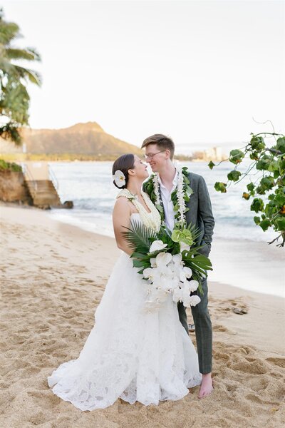 A newlywed couple standing close together as they lean in for a kiss while standing on a beach 