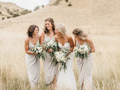 Bride in a white gown standing in a grassy field with three bridesmaids in light gray dresses, all holding white floral bouquets and smiling together. Hair done by Victoria Carling