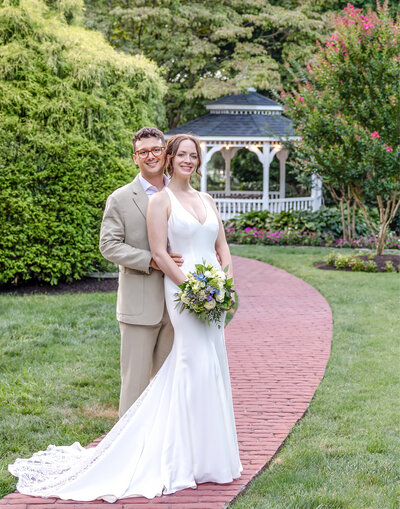 The bride and groom pose on a brick path in a lush garden with a white gazebo in the background, captured during a Wilmington, DE wedding, blending natural beauty with classic romance.
