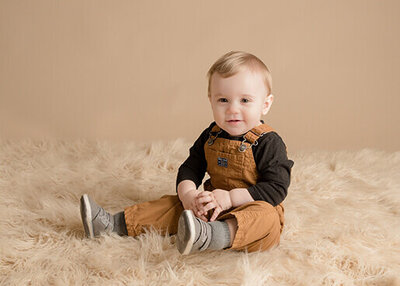 Little boy is wearing brown overalls and sitting on a tan rug while he smiles at the camera.