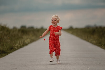 Een meisje in een rood pakje met een bloem in haar handen die lachend rent over een grijze weg met groen gras aan de zijkant