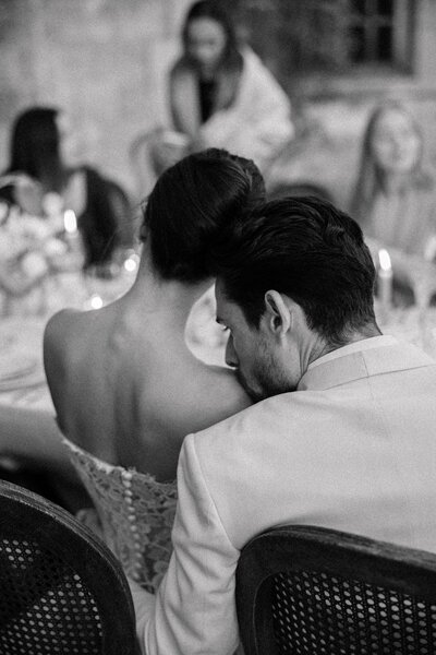 Candid black and white shot of groom kissing bride’s shoulder at the dinner table.