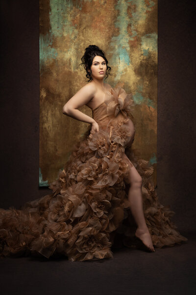 A high fashion maternity photograph with a brown and gold gown covered in large tulle flowers and a brown background with a strip of teal and gold behind the pregnant woman with her dark in an elegant updo