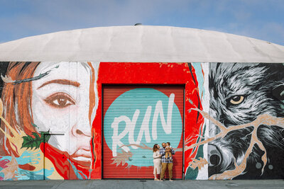 Dad holding baby in front of colorful Oakland mural with mom standing next to dad and both looking at the boy