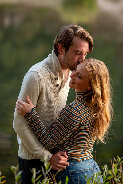 Man kisses a woman on the side of her face during their engagement session. Woman is wearing a striped shirt and the man is wearing a tan sweater.