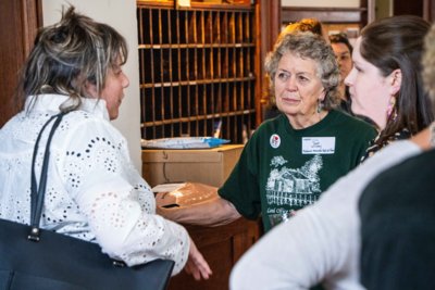 A group of people engaged in conversation at an indoor event. An older woman in a green shirt with a name tag and "I ❤️ NY" button listens intently while others stand nearby in a warm, social setting.