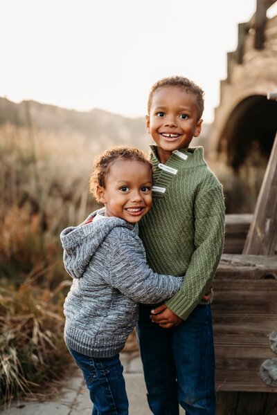2 brothers hugging. there is tall grass and the martinez waterfront bridge behind them