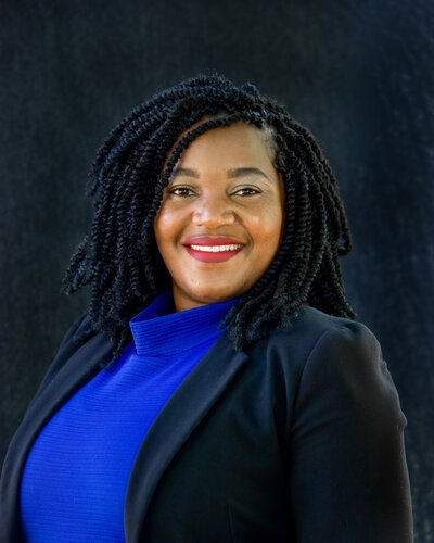 a studio branding portrait of a black woman with corn rows wearing a black blazer and bright blue mock turtleneck