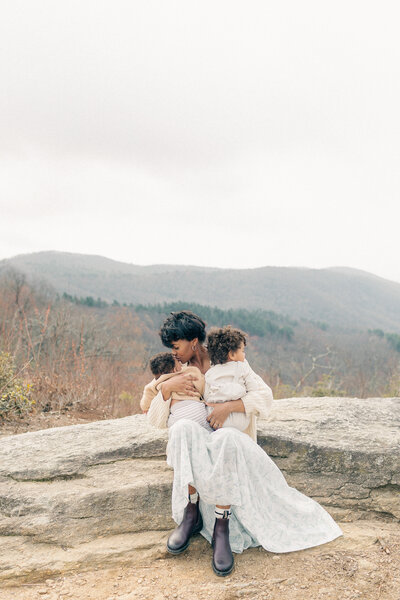 Mother in a floral dress sits on a rock holding her newborn and toddler, captured in a Kansas City family session.