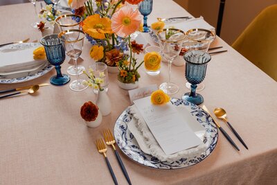 Sophisticated wedding tablescape with bright orange and yellow flowers, blue goblets, and gold-accented tableware for a refined event setting.
