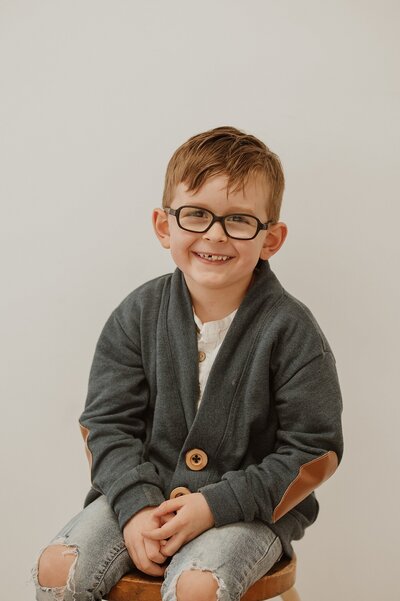 Small boy with glasses and a big smile sitting on a stool