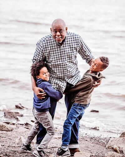Grandfather hugs his two grandsons by the Hudson River in Westchester, NY, showing a bond of love and joy.