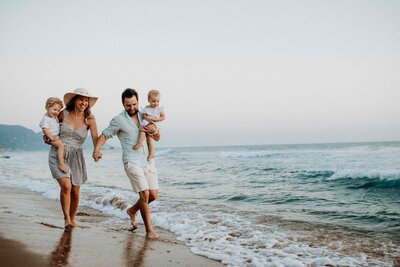 A family of four, with two adults and two young children, walks hand in hand along a sandy beach near the ocean. The sky is clear, and gentle waves are in the background. The adults are wearing casual beachwear, and the atmosphere is relaxed.