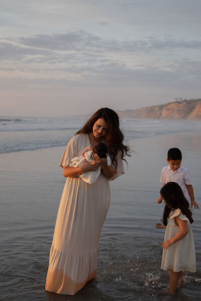 Mother holding her newborn while her two older children play at the water's edge on the beach